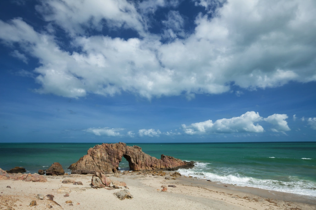 Praia com formação rochosa, mar esverdeado, areia clara e imensidão do céu ao fundo