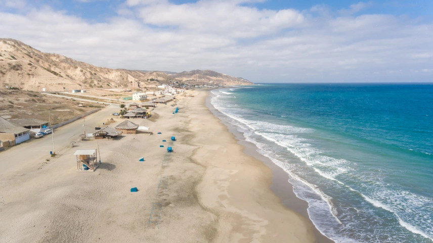 Vista aérea de uma praia com areia clara e mar azul turquesa. Ao fundo, alguns montes da mesma cor da areia