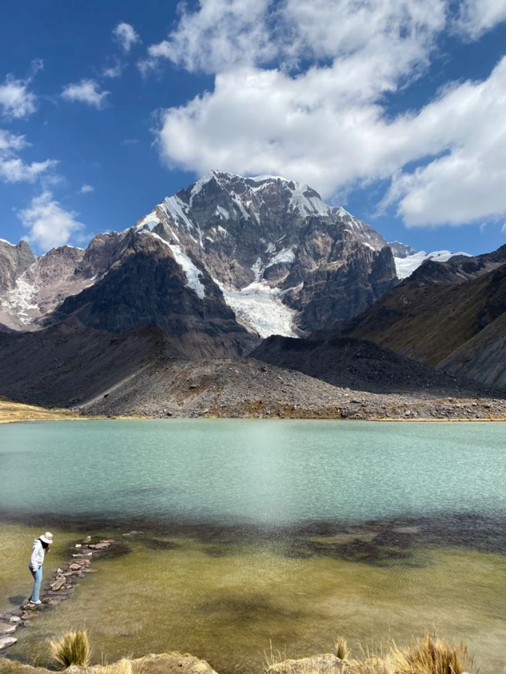 Vista panorâmica de uma lagoa azul, com uma mulher em pé sobre pedras no canto esquerdo. Ao fundo, montanhas com algumas partes cobertas por neve, na região dos Andes no Peru.