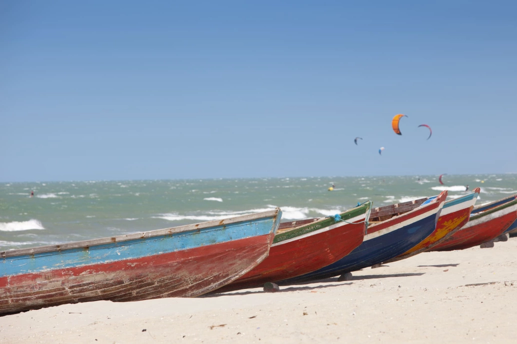 Barcos sobre a areia da praia. Ao fundo, a imensidão do mar e algumas pipas usadas na prática de kitesurf