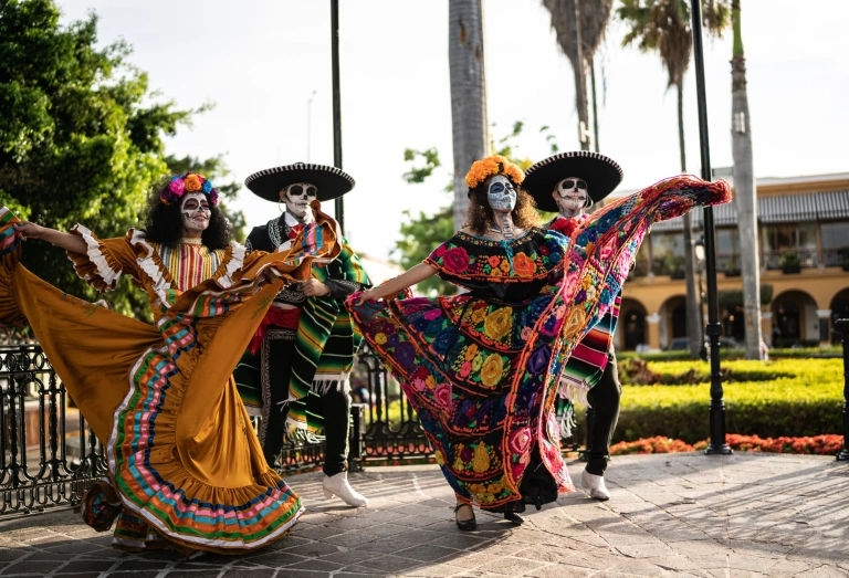 Duas mulheres e dois homens com maquiagem de caveira mexicana e trajes típicos dançando em uma praça.