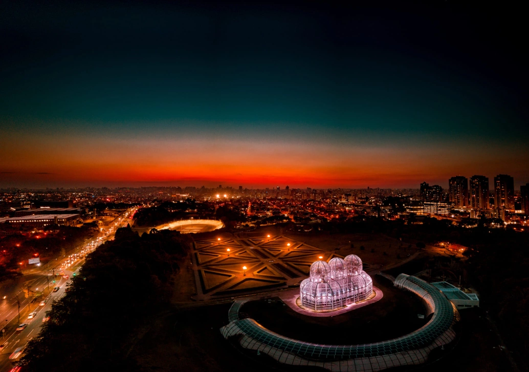 Vista aérea noturna do Jardim Botânico de Curitiba. Destaque para a cúpula de vidro iluminada. Também há luzes da cidade, e o céu está em degradê azul e laranja