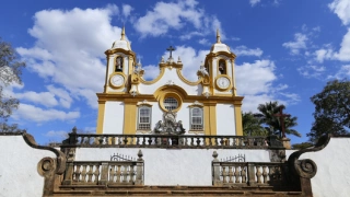 Igreja colonial em estilo barroco em cidade histórica de Minas Gerais, vista através de sua escadaria. Ao fundo, céu azul com nuvens