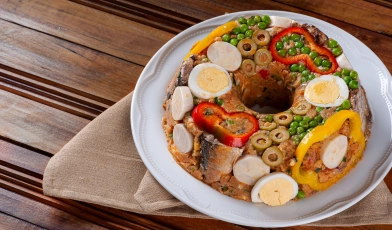Em uma mesa de madeira clara, um prato branco com cuscuz paulista, decorado com ovos, pimentões, ervilhas, sardinhas e azeitonas.