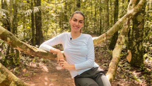 Mel Fronckowiak senta-se em galho de árvore em meio a Floresta Amazônica e sorri para a foto.