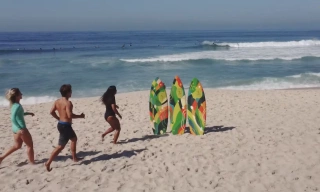Três pessoas correndo na praia em direção a três pranchas de surfe fixas na areia
