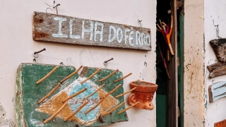 Close em uma placa com o texto Ilha do Ferro afixada na parede da entrada de uma casa junto com outros artesanatos coloridos