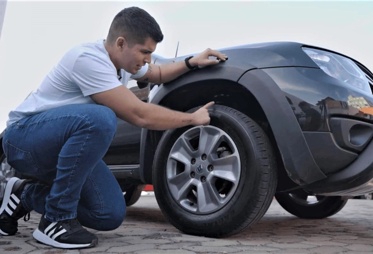 Agachado, o especialista em autos e influenciador Xenão aponta para o pneu de um carro.