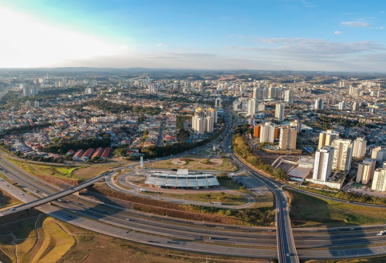 Vista aérea de uma paisagem urbana com estradas, ruas, edifícios e demais construções na cidade de Jundiaí, interior de SP. A foto ampla destaca o céu azulado e os bairros em expansão no entorno do município.