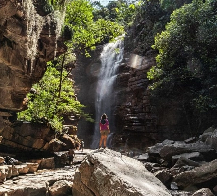 Uma mulher em pé em uma pedra contempla uma enorme cachoeira cercada por formações rochosas e vegetação nativa