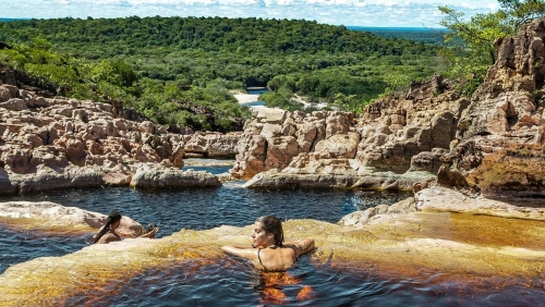 Duas pessoas nadam em piscina natural de uma cachoeira cercada por rochas e vasta vegetação preservada em dia claro