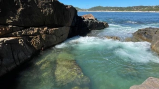 Piscina natural de água cristalina esverdeada envolta de rochas em praia catarinense. Um lindo céu azul se destaca ao fundo.