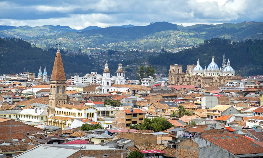 Vista aérea da cidade de Cuenca. No plano frontal, topos de casas e várias igrejas coloniais. Ao fundo, montanhas com cobertura vegetal