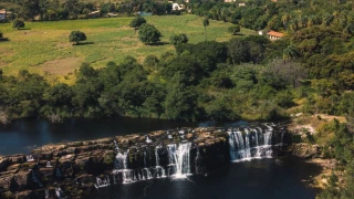 Vista aérea de cascatas sobre um paredão de pedra extenso, formando um poço natural em meio à vegetação nativa. Ao fundo, um campo plano e gramado com algumas árvores e montanhas cobertas por vegetação