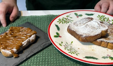 Um prato com decorações natalinas e uma pedra de ardósia servidas de fatias de pães. Uma, coberta de açúcar e doce de leite, outras duas, somente de açúcar. É conhecida como rabanada.