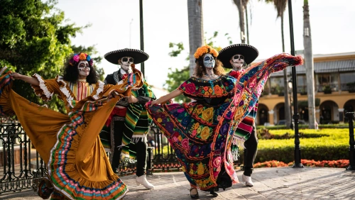 Duas mulheres e dois homens com maquiagem de caveira mexicana e trajes típicos dançando em uma praça.