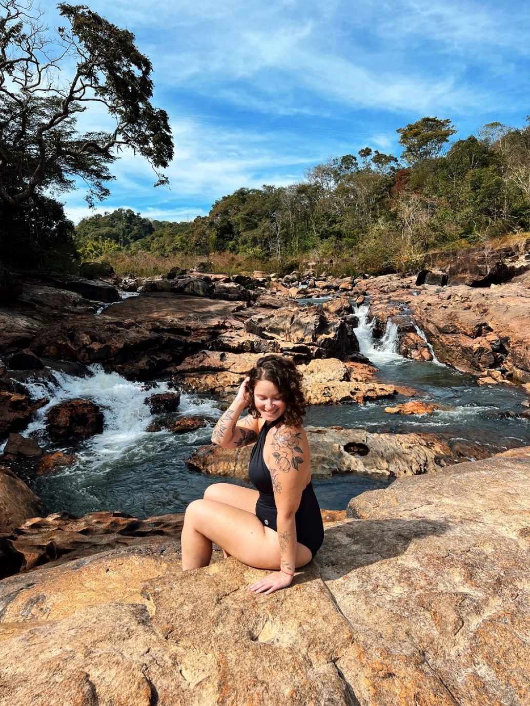 Mulher posando para foto sentada em uma pedra. Ao fundo, piscinas naturais formadas por pequenas quedas d’água