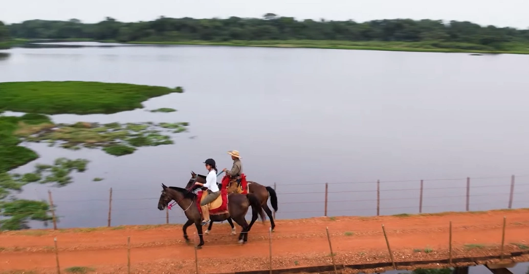 Mulher e homem cavalgam por estrada de terra vermelha cercados por rio pantaneiro