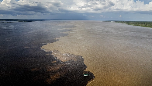 Foto apresentando fenômeno de encontro entre dois rios, o Rio Negro e o Rio Solimões. Do lado esquerdo da foto, águas escuras contrastam com águas em tom amarronzado do lado direito da foto.